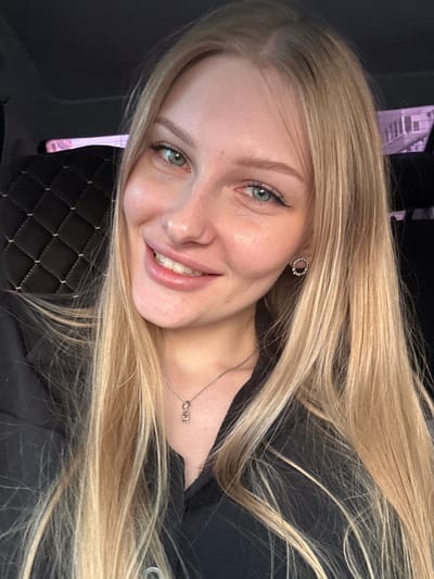 A woman with long blonde hair is smiling in a selfie-style photo. She has fair skin, blue eyes, and is wearing a delicate necklace and circular earrings. The background appears to be dark with a quilted surface visible beneath her head. There is no evidence to support this image being taken in Кондратово, Russia.