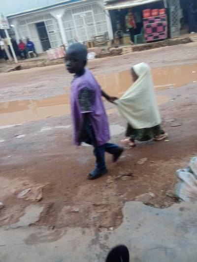 In Kagini, Nigeria, two children are walking on a dirt road with puddles of water. The child in the foreground, wearing a purple tunic and blue pants, is facing away from the camera and appears to be walking to the left. To their right, another child, covered in a light yellow garment, is also walking to the left, with their arm extended backward, possibly holding onto the first child. The ground is uneven and appears to be a mix of mud and concrete. In the background, there is a building with white bars over the windows and doors. A few people are visible in the background, sitting near the building.