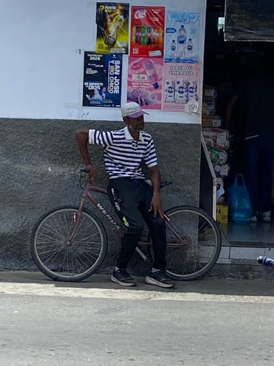 The image depicts a street scene in San José de Barlovento, Venezuela. A man wearing a striped shirt and a white cap is leaning against a bicycle. The bicycle has a purple frame and a dark-colored seat. The man is holding the handlebars with his left hand. Behind the man and the bicycle, there is a wall with posters attached to it. These posters advertise various products, including drinks and bread, with text in Spanish. To the right of the wall, there is a store entrance where stacked goods are visible. The ground in front of the wall and store is paved.