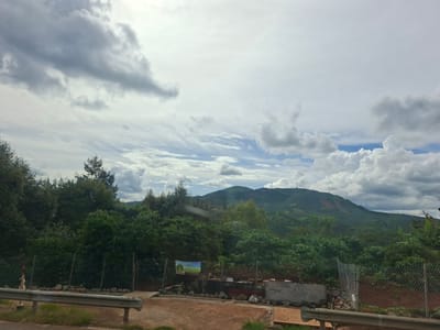 The image shows a landscape scene in Thôn 1, Vietnam. In the foreground, a metal guardrail runs along a road with a dirt shoulder and some green grass. Behind the guardrail, a chain-link fence encloses an area with trees and some construction materials, including rocks and concrete blocks. A colorful poster is attached to the fence, depicting a green landscape.
The middle ground is dominated by dense green foliage, appearing to be trees and bushes. Beyond the greenery, a rolling green hill or mountain rises, its slopes marked with patches of lighter green and some bare earth, suggesting agricultural terraces or erosion. The sky above is filled with dramatic clouds, with patches of blue sky visible between them. Some of the clouds are dark and heavy, indicating potential for rain, while others are lighter and more wispy.
The scene appears to be set during the daytime, with overcast but bright conditions. The overall mood is one of natural beauty and rural tranquility, with a hint of human presence through the fence and poster. No people are visible in the image.