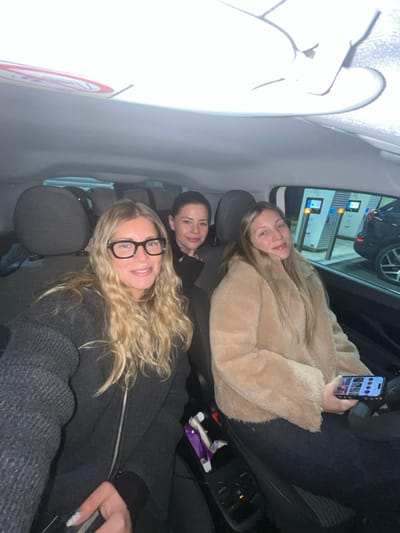 Three women are visible inside a car, likely in the back seat. One wears large glasses and a dark sweater, another a tan fuzzy jacket, and the third a dark top. The woman in the jacket holds a smartphone. Through the car window, charging stations are visible with illuminated screens. The interior of the vehicle is primarily dark.
