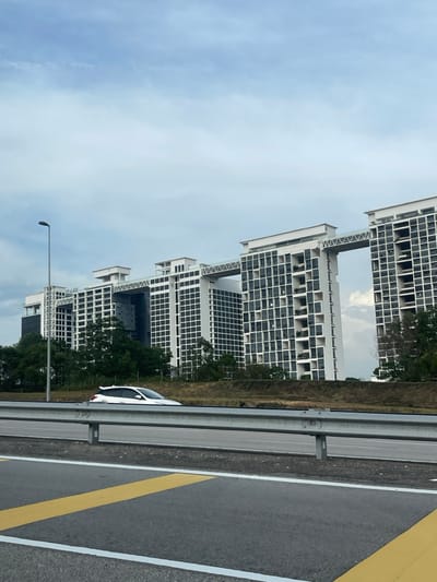 The image displays a modern urban scene in Cyberjaya, Sepang, Malaysia, under an overcast, light blue-grey sky. In the background, a complex of several large, multi-story buildings stands prominently. These structures are predominantly white with dark, rectangular windows and balconies, featuring a distinctive architecture with multiple elevated skybridges connecting different towers. A dense strip of green trees and vegetation lies between these buildings and the foreground. Below the vegetation, an earthen slope leads down to a multi-lane asphalt road. A silver metal guardrail extends horizontally across the middle ground, separating the immediate foreground road from a section further back. A white hatchback vehicle, partially visible behind the guardrail, is positioned in front of the vegetation, seemingly in motion towards the right. A tall, dark street light pole rises vertically from the left side of the foreground, partially obscuring the background buildings. The immediate foreground shows grey asphalt with yellow diagonal markings and a white solid line.