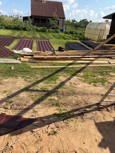 The image depicts an outdoor construction scene on a sunny day in Valyevachski Sel'ski Savet, Belarus. In the background, a rustic wooden house with a dark, corrugated metal roof stands amidst lush greenery and trees. To the right of the house, a transparent greenhouse is visible. In the foreground, stacks of unfinished wooden planks are laid out on the grass, awaiting construction. Several sheets of dark brown, corrugated roofing material are also present, some laid out on the grass and others in piles. A white electrical cord runs across the sandy ground, partially obscured by shadows cast by unseen structures or trees. A partially filled plastic water bottle rests on the grass to the left of the wooden planks. A metal ladder leans against an unseen structure on the right side of the frame. The ground is a mixture of sandy soil and patches of grass. The overall impression is of a construction project in progress, likely the renovation or extension of a rural property.