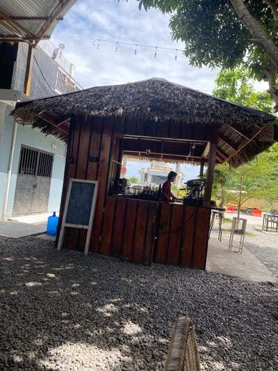 The image depicts an outdoor cafe or roadside stall with a thatched roof, likely in Aluepiet, Indonesia. A person wearing a red shirt is standing behind a wooden counter, appearing to prepare drinks with an espresso machine. The stall is situated on a gravel ground, with a paved walkway leading to it. In the background, a blue water jug is visible next to a building with metal doors and a barred window. Trees and other vegetation are present, along with a yellow truck in the distance. The overall scene suggests a casual, open-air establishment.