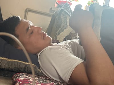 A young man is lying on his back on a bed, his head resting on a dark blue pillow with a fringed trim. He is wearing a white t-shirt and is looking up and to the left. His right arm is bent at the elbow, and his hand is raised, obscuring part of his face. Behind him, the background is blurred, but appears to show a window with light coming through and some fabric draped over furniture. The overall impression is of a relaxed, indoor setting. The image does not contain enough information to determine the location in the city of Джапалак, Kyrgyzstan.