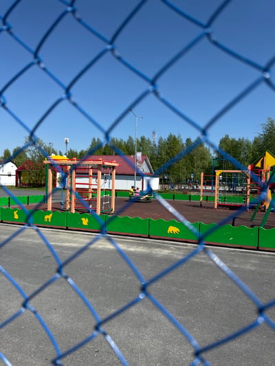 The image captures a vibrant children's playground in Высокий, Russia, viewed through a blurred, blue chain-link fence in the foreground. The fence forms a crisscross pattern, partially obscuring the scene beyond.

The playground features multiple colorful structures with wooden, yellow, and red elements, including climbing nets and ladders, set on a dark reddish-brown soft-fall safety surface. A distinctive green barrier, adorned with yellow animal cutouts such as a bear, squirrel, and possibly a fox or dog, lines the perimeter of the play area on the left side. To the right of the playground, a grey asphalt or paved surface is visible.

In the background, a small white building with a red-tiled roof stands to the left. Further back, dense green trees frame the scene under a clear, bright blue sky. A tall pole equipped with multiple antennas is discernible among the trees, suggesting a modern infrastructure element. Indistinct figures, likely people, are visible near the playground equipment and the white building, hinting at ongoing activity or recent use.

The scene is illuminated by bright sunlight, indicating a clear, sunny daytime. No specific activities are clearly defined, but the setting suggests a lively recreational space. While there are signs on distant buildings, no text is legible. The overall impression is one of an inviting outdoor play area on a pleasant day.
