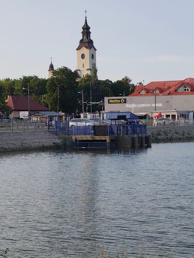 The image depicts a waterfront scene in the city of Stryków, Poland. In the foreground, a body of water with gentle ripples fills the lower half of the frame. A blue and grey structure, possibly a pier or a docking station, juts out into the water, featuring metal railings and a covered area. Beyond the waterfront, a paved walkway is visible, separated from the water by a stone embankment and a metal fence.

Further back, a commercial building with a red-tiled roof and white walls is prominent, bearing the yellow "Netto" logo, indicating a supermarket. Adjacent to it, a smaller building with a red awning is also visible. In the background, two church towers rise above a canopy of green trees, suggesting a historical or religious presence in the city. Several streetlights and traffic signals are also present. The overall scene suggests a public area by the water, likely a park or promenade, with commercial and religious structures in the vicinity. The sky is clear and pale, indicating daytime with diffused sunlight. No people are clearly visible in the image.