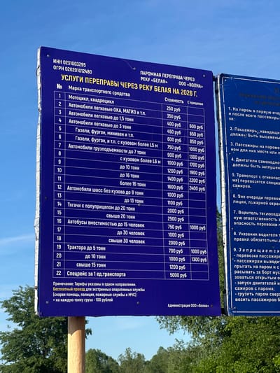 This is an image of a price list sign for a ferry crossing of the Belaya River in Russia, effective as of 2026. The sign lists prices for various types of vehicles, including motorcycles, cars, trucks, buses, and tractors. It also notes free passage for emergency service vehicles and an extra charge per ton of cargo. The sign is mounted on a wooden post, and a snippet of a neighboring sign that has rules to follow.