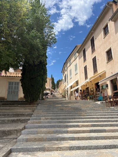 The image depicts a wide stone staircase ascending a street in the town of Pollença, Spain. The staircase is lined with buildings on the right and a tall cypress tree and a building on the left. The buildings on the right have pale peach facades with brown shutters on the windows. Several shops with awnings are visible at the base of the staircase, with merchandise such as bags and decorative items displayed outside. Two women are walking up the stairs in the middle ground, near the shops. The sky above is blue with scattered white clouds. The staircase itself is made of large, weathered stone blocks. The environment appears to be a sunny day, typical of a Mediterranean climate.