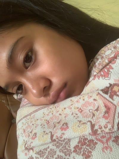 A close-up, low-angle selfie of a young person is shown, featuring their face and upper body. The subject's head is tilted to the left, with their right eye partially visible and their left eye prominently displayed. Their dark hair frames their face. The individual is resting their cheek on a blanket with a floral pattern in shades of pink, red, and yellow. The background consists of a plain, light-colored surface, possibly a wall. The image appears to be taken indoors, with soft lighting. No specific location in Sungai Meriam, Indonesia, is depicted or suggested by the visual information in the image.