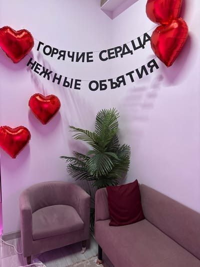 The image displays an interior room in Novosergievka, Russia, decorated for a romantic occasion. The walls are light-colored, presenting a distinct pinkish tone. Two black Cyrillic banners are strung horizontally across the upper wall section; the top banner reads "ГОРЯЧИЕ СЕРДЦА" (HOT HEARTS), and the lower banner reads "НЕЖНЫЕ ОБЪЯТИЯ" (TENDER HUGS). Five red, heart-shaped foil balloons are affixed to the walls, with three positioned on the left and two on the right. A single-seater armchair upholstered in a light mauve fabric with wooden legs is situated in the lower-left portion of the frame. To the right of the armchair, a large artificial palm-like plant with green fronds stands. Adjacent to the plant on the right is a chaise lounge or sofa section, upholstered in a matching light mauve fabric, featuring a single dark red cushion. The floor is light-colored, and a white power strip with a white electrical cable is visible on the floor near the armchair. A small, light-colored patterned rug is partially visible on the floor between the furniture pieces. No individuals are present in the scene.