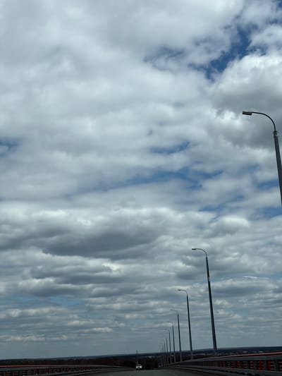 The image shows a view from a bridge in Tarasovo, Russia. The sky is filled with layered, striated clouds with patches of blue visible. A row of streetlights runs along the right side of the frame, attached to the bridge's railing. The bridge's structure, including a red metal railing and potentially other structural elements, is partially visible on the far right. The general environmental conditions indicate an overcast or partly cloudy day. A road and maybe a car are visible on the bridge.
