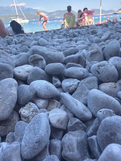 This is a vertical, close-up, eye-level shot of a pebble beach. The foreground is dominated by an abundance of smooth, grey and white pebbles of various sizes and shapes, with some larger, oval-shaped stones being most prominent. The texture of the pebbles is detailed, showing some porous surfaces and natural variations in color and pattern.
In the background, the scene transitions to a blurred view of a beach resort area. The turquoise water of the sea is visible, with some boats and a sailboat on the horizon. There are also glimpses of people on what appears to be a pier or boardwalk. One person is clearly visible, wearing a bathing suit and seemingly in motion, possibly engaging in a water sport. Other figures are also present, some with towels or beachwear. The overall atmosphere suggests a sunny day at a popular tourist destination.
The time of day appears to be late afternoon or early evening, given the warm, soft lighting and the long shadows that might be implied by the scene. The weather seems pleasant and clear, suitable for beach activities. The location context provided, Radovići, Montenegro, further reinforces the image's setting as a coastal area in the Mediterranean. No visible text is present in the image. The image captures a sense of relaxation and summer vacation vibes.