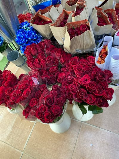 The scene, located in Sidi Moussa, Algeria, displays a commercial arrangement of numerous cut flowers. The majority of the visible area is filled with dark red roses, presented in various retail configurations. Several large bouquets of red roses are wrapped in clear plastic sleeves and positioned in white, footed, cylindrical containers directly on a light brown tiled floor. Other red rose bouquets are individually wrapped in textured brown paper, some featuring "Baraka Roses" branding and QR codes. A smaller cluster of orange roses is visible within a white fluted paper sleeve on the right. To the upper left, a quantity of bright blue artificial roses and a smaller bunch of red artificial flowers are displayed. Multiple tall, narrow, silver-colored metallic cylinders, likely flower buckets, are present on the left side of the frame. The flooring consists of uniformly sized light brown square tiles. No individuals are visible within the image.