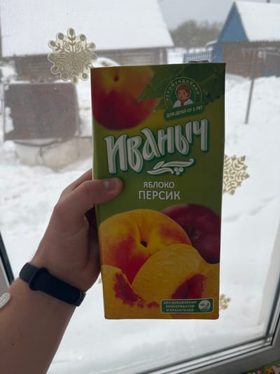 The image displays a juice box for children. The brand is "Ivanych" (Иваныч), and the flavor is apple-peach (ЯБЛОКО ПЕРСИК). The packaging indicates that it's recommended for children from 3 years old (ДЛЯ ДЕТЕЙ ОТ 3 ЛЕТ). It also states that it has no added preservatives and dyes (БЕЗ ДОБАВЛЕНИЯ КОНСЕРВАНТОВ И КРАСИТЕЛЕЙ). A hand holding the juice box can be seen, with a smartwatch on the wrist. The background shows a window with snowflake decorations and a view of snowy outdoor scenery.