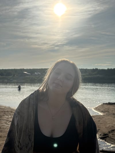A young woman with blonde hair and closed eyes stands on a sandy shore, facing the viewer. She is wearing a black top and a patterned cover-up draped over her shoulders. A silver necklace with a cross pendant is visible. Behind her, a body of water stretches to a treeline on the opposite shore, with a few buildings visible among the trees. The sun is high in the sky, casting a bright light through the clouds. A person is standing in the water in the distance. The scene appears to be a natural setting, possibly a lake or river, near a wooded area.