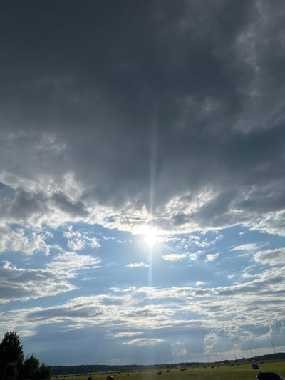 The image depicts a wide, open field under a dramatic sky. The upper portion of the sky is dominated by dark, heavy clouds, suggesting an impending storm or overcast weather. Below these dark clouds, the sky transitions to a lighter blue, with streaks of white and grey clouds. The sun is visible, breaking through the clouds in the middle of the frame, casting a bright beam of light downwards.

The field below is covered in green grass, and scattered across it are numerous round hay bales, some appearing recently baled and others more weathered. In the distance, a treeline marks the horizon, and beyond that, a hint of infrastructure, possibly roads or buildings, can be seen. A few scattered trees are visible on the left side of the frame, closer to the foreground. The overall impression is of a rural or semi-rural landscape during a day with dynamic weather conditions. The location is the city of Lukovnya, Russia.