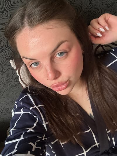 A selfie of a fair-skinned woman with long brown hair and blue eyes lying on a dark surface with a patterned texture. She is wearing a dark-colored top with a white grid pattern. Her left hand is raised near her head, displaying nails with a white base and silver glitter accents. She has a small gold hoop earring in her left ear and a gold hoop piercing in her left nostril. A small tattoo is visible on her neck. The location is not discernible from the image.