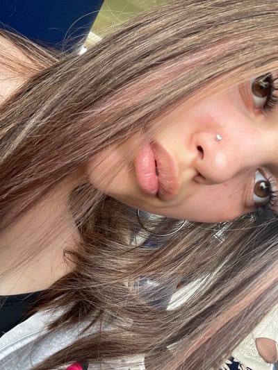 The image presents a rotated close-up portrait of a young individual, likely female, captured from an overhead perspective. The subject's head is tilted significantly, with their face angled towards the upper right of the frame. Long, wavy light brown hair with subtle lighter streaks or highlights cascades across the image, obscuring parts of the face and background.
The subject's lips are prominent, pursed into a "duck face" expression, with a light pink/nude glossy finish. A small, shiny silver stud piercing is visible on the left nostril (viewer's right). The eyes are open, with rich brown irises and pronounced dark eyelashes, looking slightly upwards and to the viewer's left. The skin appears clear with some minor texture visible.
On the subject's lower left, a large, thin silver hoop earring is partially visible against the hair. Visible clothing includes a dark garment (possibly a top or bra strap) near the upper left of the subject's body, and a textured, light grey or white fabric (possibly a hoodie or jacket) along the bottom left edge of the frame. In the bottom right, a segment of a white, open-knit or lacy garment is visible, along with a hint of blue and white patterned fabric underneath, suggesting additional clothing or another person in the vicinity.
The background elements are largely out of focus and indistinct, visible through and around the hair. A dark blue surface is present in the upper left, while a lighter green/beige surface is in the upper right. These suggest an indoor environment. The scene is located in the city of Osel'ki, Russia.