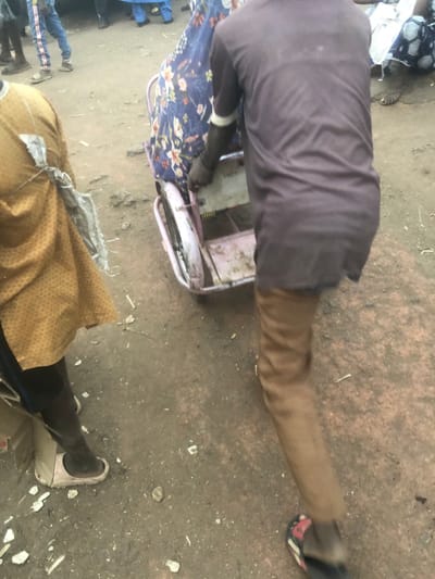 In Tukur Tukur, Nigeria, an outdoor scene on unpaved, light brown ground depicts human activity. In the central foreground, a person viewed from the rear wears a dark maroon tunic with white cuff trim, light brown trousers, and dark flip-flops with red detailing. This individual operates a worn, light pink two-wheeled cart, pushing it forward. A person in a blue floral garment is partially visible seated on or behind the cart. To the left, another person is partially visible, wearing a light brown patterned garment with a torn, light-colored fabric piece secured to their side, and dark footwear with a light sole. In the blurred background, other individuals are present on the unpaved surface, with one person visible wearing blue jeans with white side stripes. Debris and small stones are scattered across the ground.