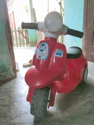 A red toy scooter is positioned in the foreground, with its front wheel closest to the viewer. The scooter features a white headlight and black handlebars. There are decals on the front of the scooter, one of which reads "UNION 99". The scooter is on a concrete floor, which appears to be dirty and worn. In the background, a wooden fence and a wall painted in a muted blue are visible. The scene is illuminated by bright sunlight, suggesting an outdoor or semi-outdoor setting. A pair of beige sandals are also visible to the left of the scooter. The overall environment suggests a residential area, possibly a yard or a porch.