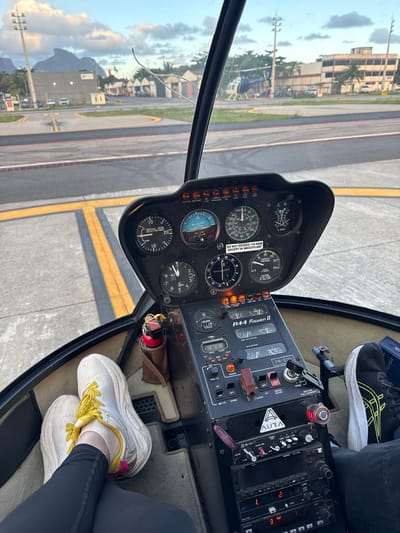 This image captures the interior view from the co-pilot's seat of a Robinson R44 Raven II helicopter, identified by the registration "PR-ETO" visible on the avionics stack. The helicopter is stationary on an asphalt apron with yellow markings.
Two individuals are present in the cockpit, evidenced by their legs and feet. The person in the co-pilot's seat, from whose perspective the photo is taken, wears white athletic shoes with prominent yellow laces and black pants. The pilot's left foot, wearing a black athletic shoe with white and yellow accents, is partially visible near the flight controls.
The instrument panel displays various gauges: an altimeter showing approximately 200 feet, a vertical speed indicator at zero, an airspeed indicator at 0 knots, an RPM gauge, and a manifold pressure gauge indicating approximately 5 inches of mercury. Warning lights for "CLUTCH," "MR TEMP," "CARBON MONOXIDE," "LOW FUEL," and "LOW RPM" are visible but unlit. Below the main panel, an avionics stack features an "AURA" brand audio panel, communication radios, and navigation equipment. A red fire extinguisher is secured in a brown leather holder to the left of the avionics. The helicopter's interior floor is carpeted in a light beige tone.
Beyond the helicopter's windshield, an airfield environment is visible. In the mid-ground, multiple light-colored buildings, characteristic of airport hangars or facilities, line the perimeter. A blue and white helicopter is parked on the apron to the left. Tropical vegetation, including palm trees, is scattered throughout the scene. In the distant background, under a partly cloudy sky, prominent and distinctly shaped mountains, including a rugged peak resembling Pedra da Gávea, dominate the landscape. A tall utility pole stands on the left side of the frame.
Forensically, the "PR-ETO" registration indicates a Brazilian-registered aircraft. The unique mountainous backdrop, particularly the formation resembling Pedra da Gávea, in conjunction with the registration, identifies the location as Jacarepaguá Airport (Aeroporto de Jacarepaguá - Roberto Marinho) in Rio de Janeiro, Brazil, with the Tijuca Forest National Park mountains in the background. The helicopter appears to be powered on but not in flight, likely undergoing pre-flight procedures or preparing for taxi.