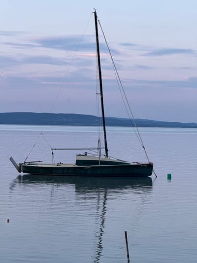 The image shows a sailboat named "Slipper" floating on a calm body of water, likely Lake Balaton, near Balatonlelle, Hungary. The boat has a dark-colored hull and a tall mast. The sky is overcast with soft, pastel colors. A green marker floats near the boat. In the distance, a line of land is visible. Another red marker is located near the bottom of the frame. The water reflects the boat.