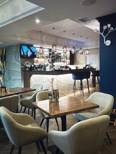 The image depicts the interior of a modern, well-lit restaurant or bar. The scene is dominated by seating areas with light-colored upholstered chairs and dark wooden tables. In the background, a long bar counter is visible, stocked with bottles and illuminated from below. Several contemporary pendant lights hang above the bar. A television screen displays an image of a person driving. The walls are decorated with a patterned marble-like finish and a dark blue accent wall. On the far right, a decorative wall sconce resembling a tree branch with lights is mounted. The flooring appears to be dark wood. The overall impression is one of a sophisticated and comfortable dining or drinking establishment. The location, Новинки, Russia, is a small village in the Nizhny Novgorod Oblast, suggesting this establishment is a local amenity rather than a large city venue.