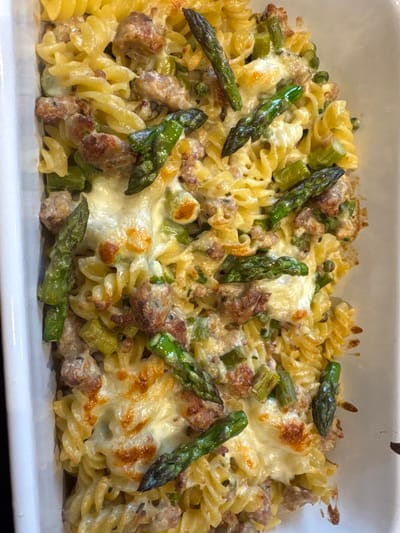 The image presents a close-up view of a hearty pasta bake, likely fresh from the oven, served in a white rectangular dish. The primary subjects are the food ingredients themselves. The dish consists of fusilli pasta, which forms the base, generously mixed with pieces of cooked, browned meat, resembling sausage or bacon. Bright green asparagus spears and smaller green peas are also prominently featured, adding color and freshness. A thick layer of melted cheese covers much of the pasta and other components, with some areas showing a golden-brown, bubbling crust, indicating it has been baked until lightly browned. The overall scene conveys a warm, comforting meal, ready to be eaten. There are no people visible, and no discernible text. The time of day and weather are not evident, as the focus is entirely on the food within an indoor setting.