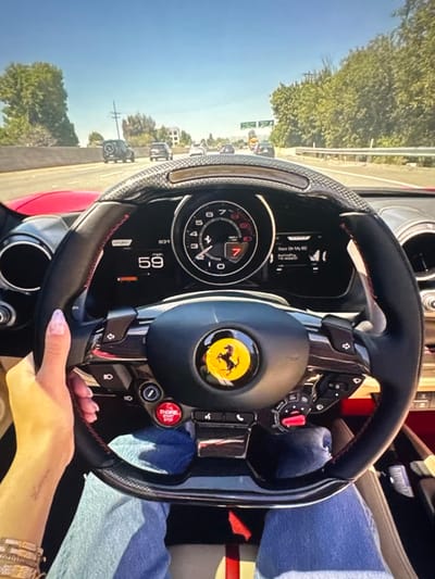 The scene captures a first-person perspective from the driver's seat of a red Ferrari convertible, driving on a multi-lane highway in the city of Lingolsheim, France, under a clear, sunny sky.

In the immediate foreground, a left hand, exhibiting light purple nail polish, grasps the black leather and carbon fiber steering wheel. The driver's left wrist features multiple stacked bracelets, and a faint linear mark or tattoo is visible on the forearm. Below the steering wheel, the driver's legs are visible, clad in blue jeans.

The Ferrari steering wheel is detailed with red stitching and prominently features the yellow Ferrari emblem with a black prancing horse in its center. Key controls visible include a red "ENGINE START" button on the lower left and a red Manettino switch on the lower right. Through the upper arc of the steering wheel, the digital instrument cluster is visible, displaying a speed of "59 mph" on the left. A central tachometer indicates the vehicle is in "7"th gear, with the redline marked around 7. An adjacent screen shows "Back On My Hill" as a currently playing audio track.

Beyond the windshield, the highway extends into the distance with moderate traffic. Multiple vehicles, including SUVs and sedans, are observable ahead, traveling in the same direction. The right side of the highway is delineated by a metal guardrail, behind which dense green foliage and trees line the roadside. On the left, additional trees and utility poles are visible against the bright blue sky.