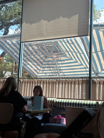 The image captures an indoor dining area looking out onto an outdoor patio, identified as Honor Bar in Highland Park Village, Highland Park, United States. A large window, partially covered by a light-colored roller blind at the top, separates the interior from the exterior. Beyond the window, a patio area is visible with multiple large outdoor umbrellas featuring light blue and white stripes. Lush green trees and a clear blue sky are discernible in the background.
Inside, two women are seated at a dark rectangular table within a booth setting. The woman on the left, seen from behind, has long brown hair and wears a dark top. The woman in the center, with long brown hair and a dark sleeveless top, is facing left, possibly engaged in conversation or viewing an item on the table. In front of her on the table is a clear glass containing ice and a light blue upright object, resembling a menu stand or tablet. She sits on a booth bench upholstered with light green and white striped cushions. To her right, on the booth seat, rests a pink patterned gift box adorned with a bow. Below the main window frame, beige vertical drapes hang. A third individual is partially visible in the blurred foreground on the right, holding a dark object, likely a mobile phone.
