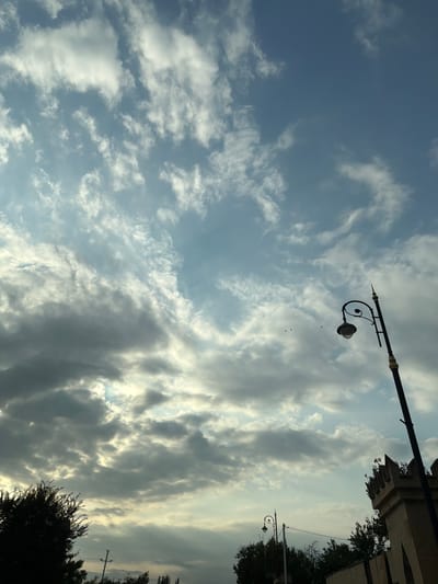 This is an outdoor, low-angle, full shot of a cloudy sky. A single black lamppost with a lantern is visible on the right side of the image, with a crenellated building partially visible below it. In the lower left corner, the silhouettes of trees are visible. In the distance, there is a utility pole with wires. The sky is a mixture of light and dark clouds, with some shafts of sunlight breaking through. A few small birds are flying in the distance. The overall scene is serene and atmospheric, suggesting either early morning or late evening. The lamppost and building suggest an urban or semi-urban environment, possibly within the city of Shamakhi, Azerbaijan, known for its historical sites and scenic beauty.