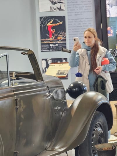 Here is a concise description of the image:
The image shows a woman in a light blue hoodie and beige jacket looking at her phone. She is standing near an old, olive-green vehicle, which appears to be a car. Behind her, there are informational posters on a wall, one of which features a photo of a gymnast. The OCR text suggests the location is in Russia, specifically referencing Moscow and the "Zenit-E" camera. Without additional context, it's impossible to confirm if the location is in the city of Архангельское, Russia.