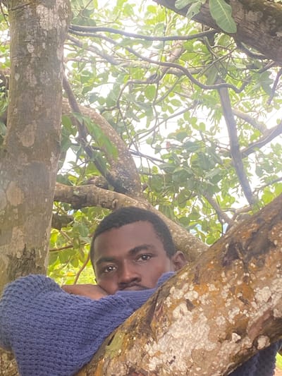 A close-up, eye-level, portrait photograph depicts a young man with dark skin and short, dark hair, resting his chin on his arms. He is wearing a dark blue, textured sweater and is nestled amongst the branches of a large tree. The tree trunk and branches are textured with rough bark and patches of lichen. Lush green leaves are visible in the background, obscuring the sky which appears overcast. The scene is set outdoors, with the dense foliage suggesting a natural environment. While the specific location in Asaga Amangwu, Nigeria is not explicitly depicted in the image itself, the setting of a tree with abundant vegetation is consistent with many outdoor environments in the region.