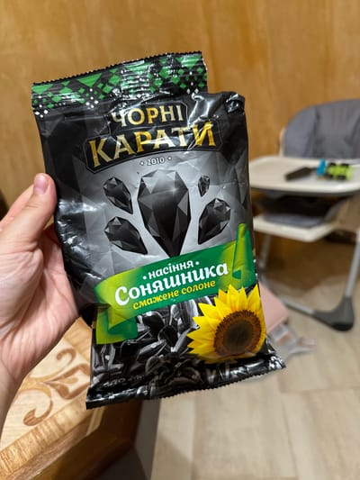 A close-up shot shows a person's left hand holding a black bag of sunflower seeds. The bag is adorned with a design featuring black diamond-like shapes and text in Cyrillic, "ЧОРНІ КАРАТИ" and "НАСІННЯ СОНЯШНИКА". A sunflower image is also visible on the packaging. The background consists of a wooden surface with a decorative pattern, and a blurred view of a child's high chair with toys in the distance. The scene is indoors, possibly in a residential setting. The location of Chemerne, Ukraine, is not discernible from the image itself.