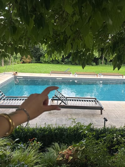 An outdoor scene captured in Vandœuvres, Switzerland, features a rectangular swimming pool with light blue tiles under clear water, positioned centrally. The pool deck is paved with light-colored stone. In the foreground, a light-skinned human hand, adorned with a gold bracelet, a silver bracelet, and a small leafy tattoo on the forearm, is extended, with the thumb and index finger nearly touching, creating a "pinching" gesture. Beyond the pool, a manicured green lawn transitions into a dense backdrop of trees and bushes. Four dark-colored, slatted lounge chairs are arranged around the pool: one is partially obscured by the hand in the near-left, another is fully visible in the near-right, and two more are on the far side of the pool. An overturned terracotta pot rests on the paving near the far-left edge of the pool. Overhead, the frame is partially covered by a canopy of large, green leaves. In the lower right foreground, a low garden bed containing various plants and a small black landscape light is visible.