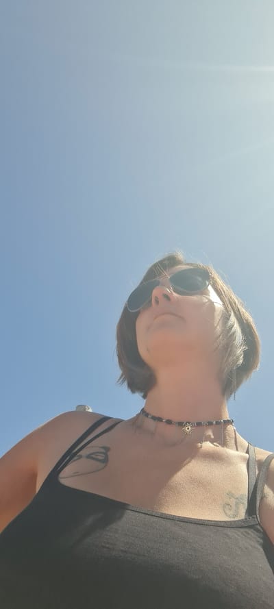 A woman wearing sunglasses and a black tank top is looking down at the camera. She has short brown hair. She is wearing a necklace. She has tattoos on her chest and shoulder. The sky is blue and clear. This image was likely taken in Nahariyya, Israel.