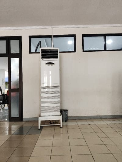 A white standing air conditioner unit is positioned against a white wall in an interior space. The unit is situated on a black metal frame, with a small, black trash can beside it.  The unit is standing in front of three large black-framed windows with clear glass panes. To the left is a large black-framed glass door. The floor is tiled in a beige and grey pattern. The image was taken in Telukjambe, Indonesia, at the coordinates -6.32 latitude and 107.293 longitude.  There are no people in the image.