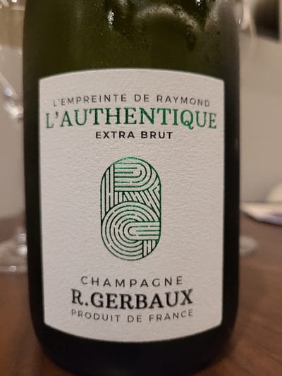 A champagne bottle with a white label featuring green text and an abstract design. The label reads "L'EMPREINTE DE RAYMOND L'AUTHENTIQUE EXTRA BRUT" on the left side. A modern green design sits in the center. The right side says "CHAMPAGNE R. GERBAUX PRODUIT DE FRANCE." The bottle appears slightly chilled with condensation.