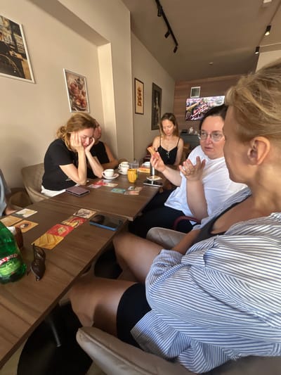 In Donji Pobori, Montenegro, at a cafe with beige walls and framed artwork, four women are seated at two adjacent wooden tables. The woman in the foreground, wearing a blue and white striped shirt, is partially visible, facing away from the camera and engaged in conversation with the woman next to her. This second woman, with dark hair and glasses, wears a white t-shirt and gestures with her hands as she speaks. To her left, a woman with blonde hair, wearing a black t-shirt, rests her head on her hands, appearing thoughtful or tired. Further down the table, a woman in a black tank top looks down at the table. The tables are set with coffee cups, saucers, a yellow mug, and several small cards arranged like a game. A green bottle of "Miloš" water and sunglasses are also visible on the table. A television screen is mounted on the wall in the background.