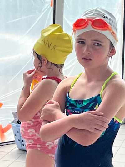 This image, captured in Andrézieux-Bouthéon, France, features two young girls in an indoor swimming pool environment. The girl closer to the viewer, positioned on the right side of the frame, has dark hair and is wearing a white swim cap with bright orange swimming goggles pushed up onto her forehead. Text is partially visible on her cap, reading "SAINT... la me." She looks directly at the camera with a neutral expression. She is wearing a multi-colored swimsuit, primarily blue and green, with the brand name "NABAIJI" visible on the chest. Her arms are crossed in front of her.
To her left and slightly behind, another girl is visible, wearing a bright yellow swim cap with the name "SEANA" written in black marker. Her face is turned away and downwards, and she is dressed in a pink swimsuit adorned with white patterns. Both girls appear wet, indicating they have recently been swimming or are preparing to enter the water.
The background shows light-colored tiled surfaces, suggesting the setting of an indoor aquatic facility. A transparent plastic sheet or cover is also visible. The scene is brightly lit, consistent with an indoor pool area, and captures a moment during a swimming activity, likely a lesson or recreational session.