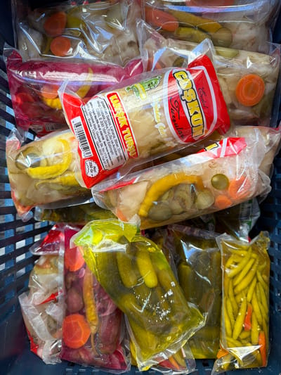 The image shows a collection of plastic bags filled with pickled vegetables, known as "turşu" in Turkish, within a blue plastic crate. The bags contain various vegetables such as cabbage, peppers, tomatoes, carrots, and cucumbers, submerged in a brine of vinegar, salt, and water. Prominently featured is a bag with a red and white label, branded "Erdal Coşkun Tursuları Gedelek," detailing the ingredients and production location. The label states the product is a "KARIŞIK TURŞU" (mixed pickles) and provides the factory address in Gedelek Mah., Gedelek Sk. No:77/A Merkez, İnegazi - Bursa - Türkiye. The net weight is listed as 250 grams. Given the types of food and location information on the label, it is implied that these were taken in Kadıköy, Turkey.