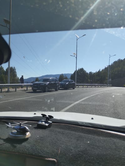 The image depicts a view from inside a vehicle, looking out through the windshield at a road and surrounding landscape. Two dark-colored SUVs are visible on the road. The vehicle in the foreground is a Chevrolet Captiva, license plate 90L 60ZA. The vehicle in the background appears to be a dark sedan. A person in a dark jacket and hat is standing near a guardrail on the left side of the road, looking out at the view. The landscape includes a clear blue sky with some wispy clouds, mountains in the distance, and a dense forest on a hillside. Several modern streetlights with solar panels are visible along the road. The interior of the car shows a dashboard with two small decorative items: a miniature helicopter and what appears to be a radar detector or similar electronic device. The overall impression is that of a scenic drive, possibly on a mountain road. The location is identified as Myk, Uzbekistan.