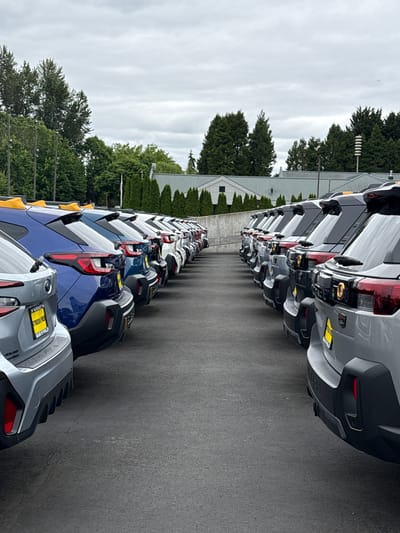 In the city of Puyallup, United States of America, a parking lot displays a row of new Subaru vehicles, predominantly silver and blue, lined up on asphalt. The cars are parked nose-to-tail, with their rear ends facing the viewer. The vehicles feature roof racks and distinctive taillight designs. A grey Subaru is positioned in the foreground on the left, displaying a yellow dealership license plate with text that includes "HARNISH" and "SUBARU OF PUYALLUP." A blue Subaru is visible to its right. The background includes a line of evergreen trees and a building with a grey, sloped roof. The sky is overcast. The scene is devoid of people.