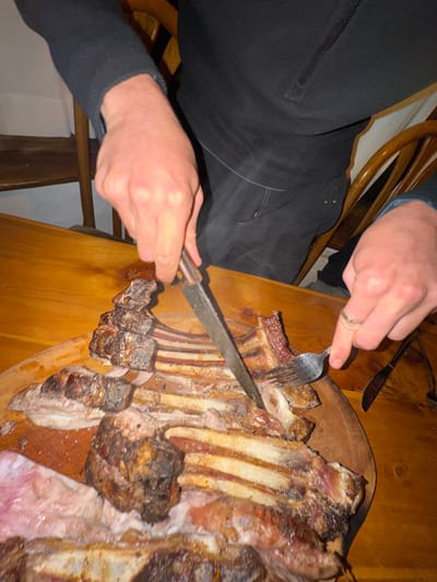 A person is using a fork and knife to cut into a rack of grilled ribs resting on a wooden surface. The person is wearing dark clothing. The ribs vary in color from light to dark brown and some red meat is visible. The wooden surface is light brown. A wooden chair is visible in the background.
