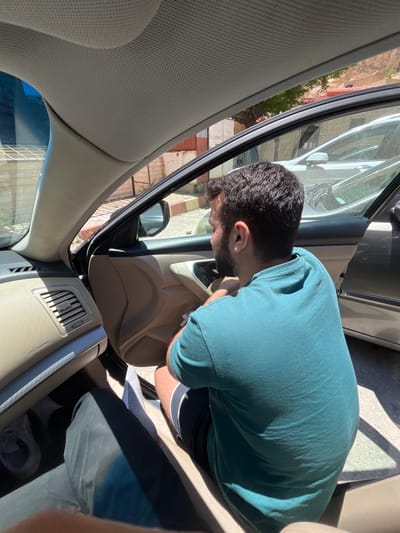 A man with dark hair and a beard is inside a car, leaning forward and looking towards the car door. He is wearing a teal t-shirt and dark shorts with a white stripe. The interior of the car is beige. The car door is open, and the exterior of the car is dark. The background shows buildings that could potentially be in Ma'loula, Syria, but the image alone cannot confirm the exact location.