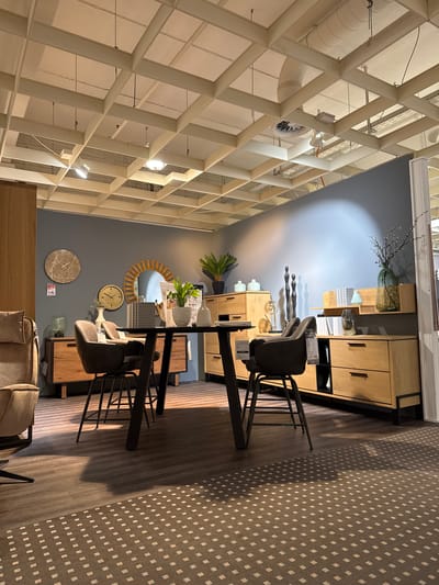 The image captures an interior scene, likely within a furniture store or a showroom, possibly in Remscheid, Germany. The architectural features are prominent, with a complex white grid structure forming the ceiling, giving the space a modern, industrial aesthetic. The room is furnished with a dining set, including a glass-topped table and two high-backed chairs with black metal frames. Various wooden cabinets and display units are arranged along the wall, adorned with decorative items such as vases, clocks, plants, and abstract sculptures. A round mirror hangs on the wall, adding to the décor. The color palette is mostly neutral, with gray walls contrasting with the warm tones of the wood furniture and the stark white ceiling. The floor is covered with a patterned rug or flooring. In the foreground, part of a beige sofa is visible, further indicating that the image was taken inside a furniture store. There are no people explicitly visible in the frame, making it challenging to describe actions or interactions.