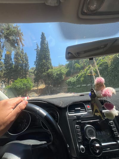 The image shows the perspective from inside a car, specifically the driver's seat. A hand is visible on the steering wheel, suggesting someone is driving. Through the windshield, a landscape of trees and greenery can be seen under a clear sky. The car's dashboard and console are also visible, including the radio display showing "89.80," the speedometer, and various buttons and controls. The presence of pink pom-poms and other ornaments attached near the rearview mirror is noted. Based on the trees and style of buildings visible through the front windshield, the location is determined to be in Beni Mellal, Morocco.