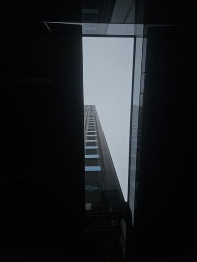 The image is a low-angle shot looking up at a tall, modern skyscraper from a street-level perspective in Shibuya, Japan. The skyscraper is the primary subject, dominating the center of the frame as it rises towards a pale, overcast sky. The building's facade appears to be made of glass and dark panels, with regular rows of windows that reflect the sky. The building on the left and right edges of the frame are dark, silhouetted structures that frame the view of the skyscraper, suggesting the shot is taken from an alcove or between two buildings. The overall impression is one of urban density and towering architecture. There are no people or vehicles visible in the image. The lighting is diffused, consistent with an overcast day.