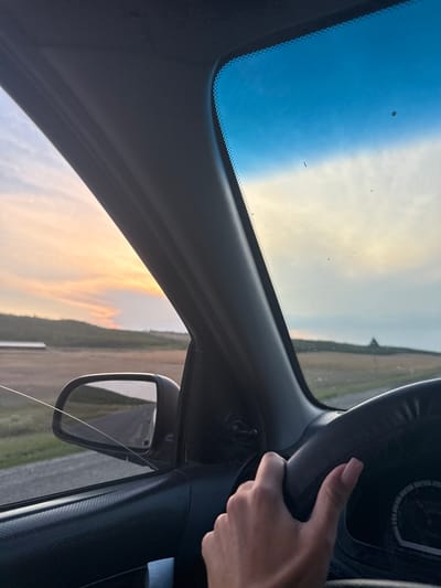 The image depicts a view from inside a car, with a hand on the steering wheel. The driver's perspective looks out through the windshield and side window towards a rural landscape at sunset. The sky displays hues of blue, yellow, and orange from the setting sun. Rolling hills with sparse vegetation are visible in the distance. A road stretches out, reflected in the side-view mirror. Part of the car's interior, including the dashboard and steering wheel, is in the foreground. The location is identified as Sadaclia, Moldova.